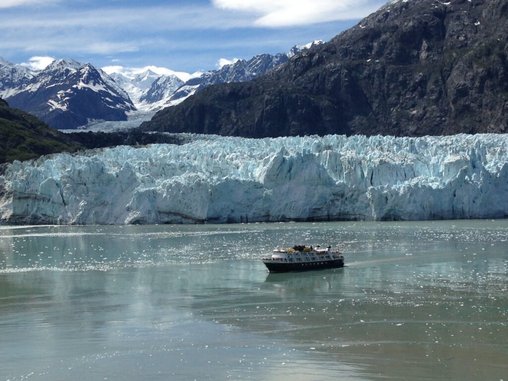 Tour boat passing a tidewater glacier in Glacier Bay, Alaska, with snow-capped mountains behind.