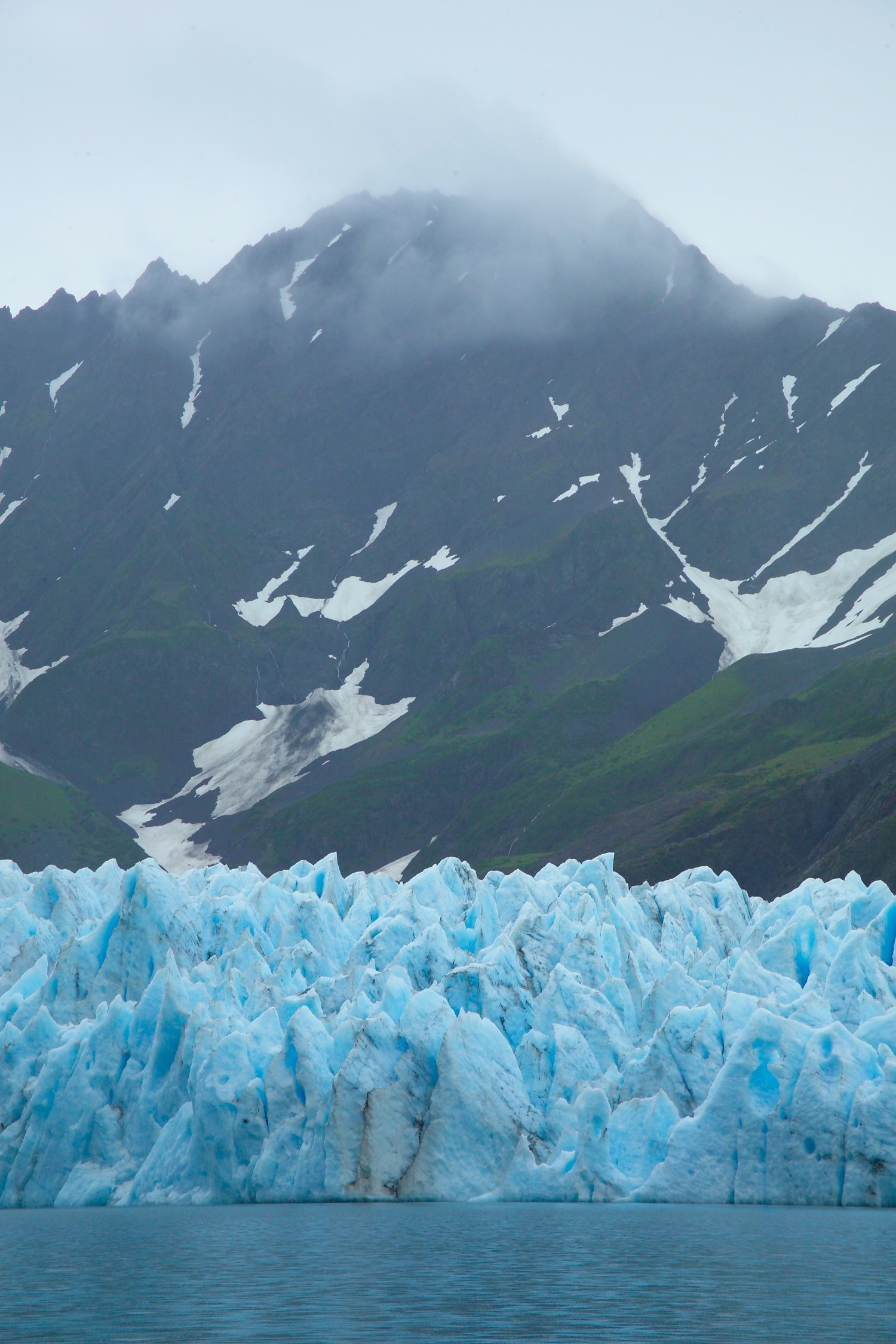 The crevassed blue-ice terminus of Pederson Glacier meeting the water below a cloud-draped mountain in Kenai Fjords National Park, Alaska.